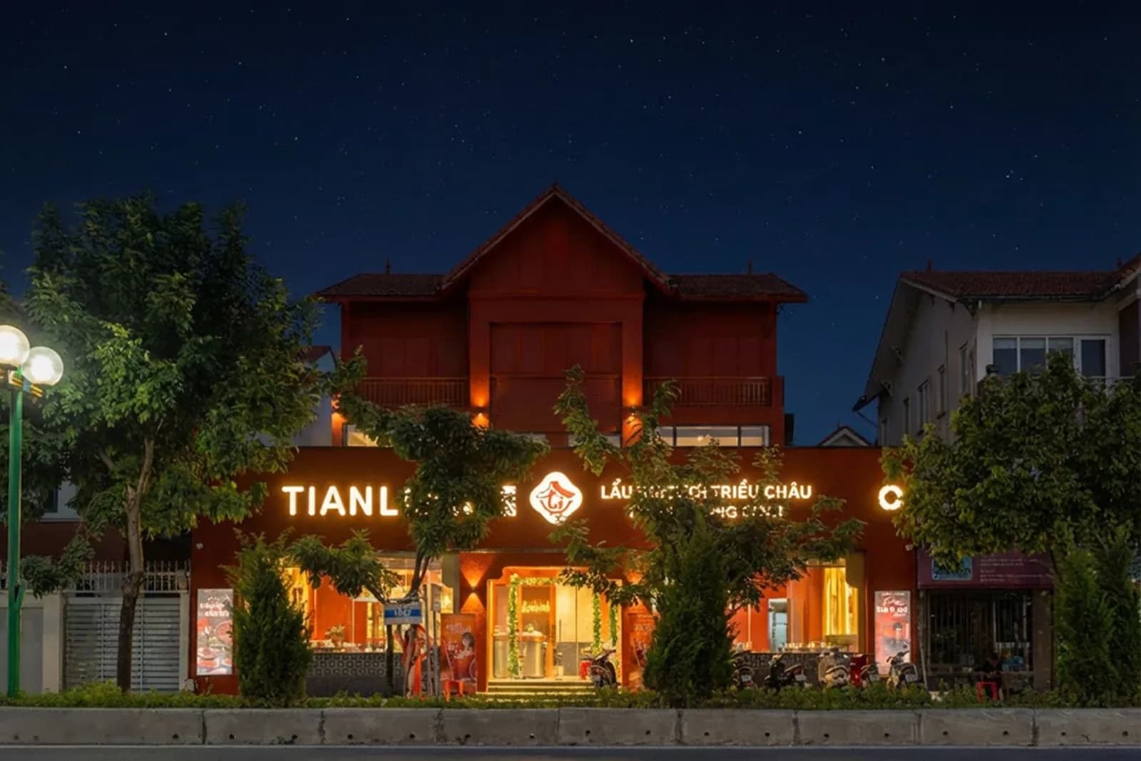 Mặt tiền Tian Long Vũ Trọng Khánh buổi tối: dải chữ TIANLONG và "LẨU BÒ TƯƠI TRIỀU CHÂU" sáng đèn trên khối 1 tầng phía trước, đèn hắt dưới ban công căn nhà mái ngói, hàng cây và cột đèn đường trước cửa.