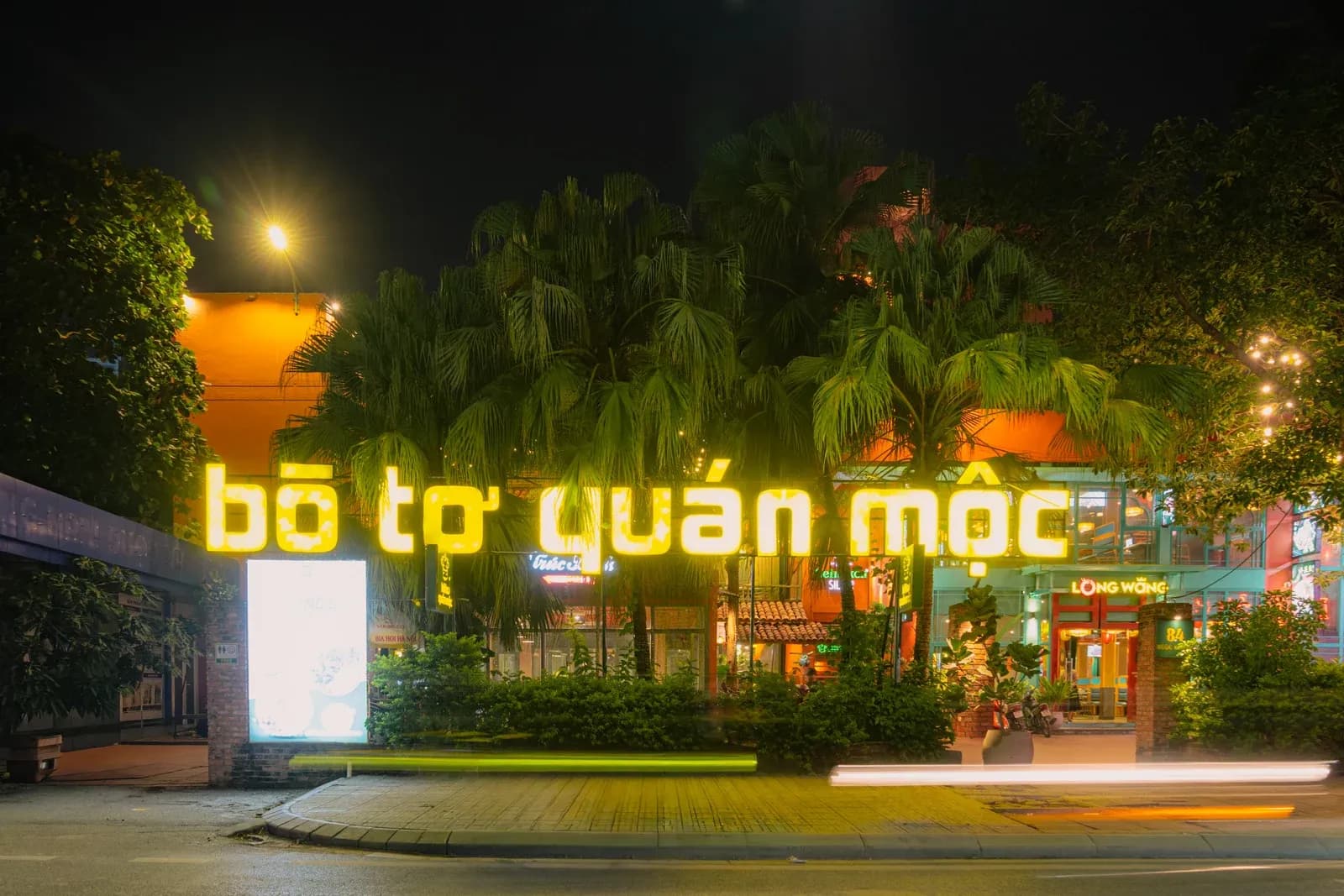 Mặt tiền quán về đêm nhìn từ lòng đường: chữ tên quán bằng đèn màu vàng nổi giữa tán cây, phía sau là khối nhà sáng đèn và vỉa hè.