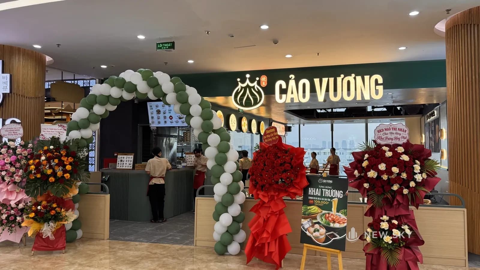 Mặt tiền Cảo Vương Vincom Ocean Park 1 nhìn thẳng từ hành lang trung tâm thương mại: biển chữ nổi phát sáng trên vách xanh rêu, quầy thu ngân bên trái, cổng bóng bay và các lẵng hoa chúc mừng đặt trước gian.