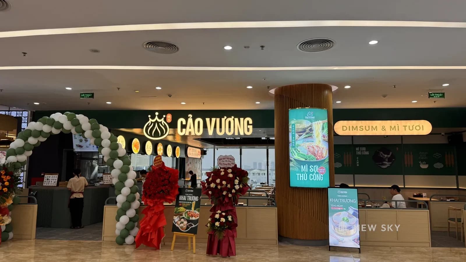 Mặt tiền Cảo Vương Vincom Ocean Park 1 nhìn từ hành lang trung tâm thương mại: biển chữ nổi phát sáng trên vách xanh rêu, cột tròn ốp nan gỗ gắn màn hình, cổng bóng bay và hai lẵng hoa chúc mừng đặt trước gian.