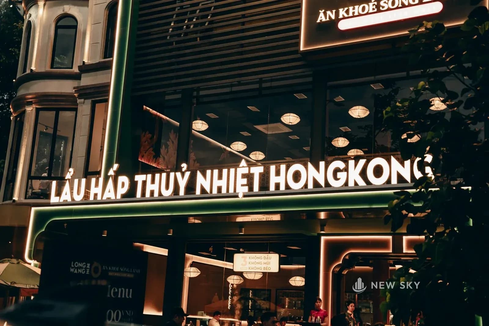 Hàng chữ nổi màu trắng ghi tên món chạy hết bề ngang căn nhà, gắn trên đầu tầng một; phía sau là dải nan gỗ nằm ngang của tầng hai và mảng kính tầng một sáng đèn.
