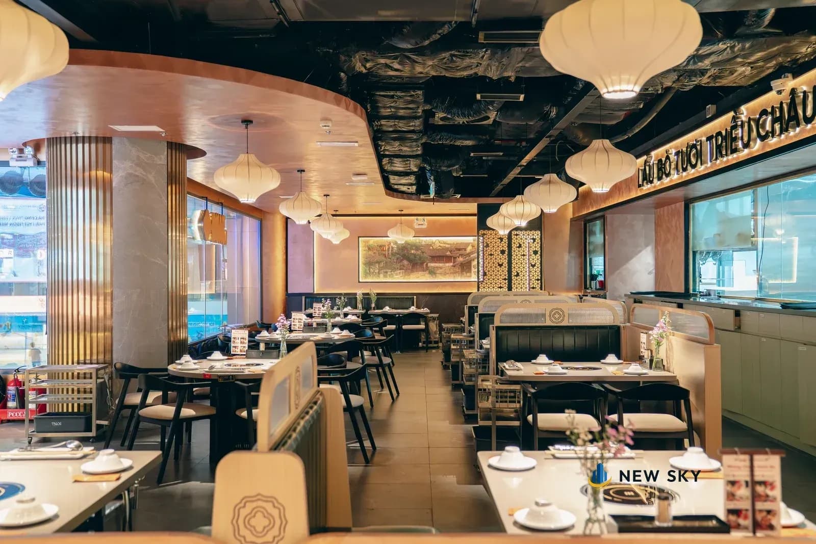Một dải trần cong màu đồng vắt ngang khoảng trần để hở sơn đen; bên dưới là hàng đèn lồng vải màu ngà và các dãy bàn.