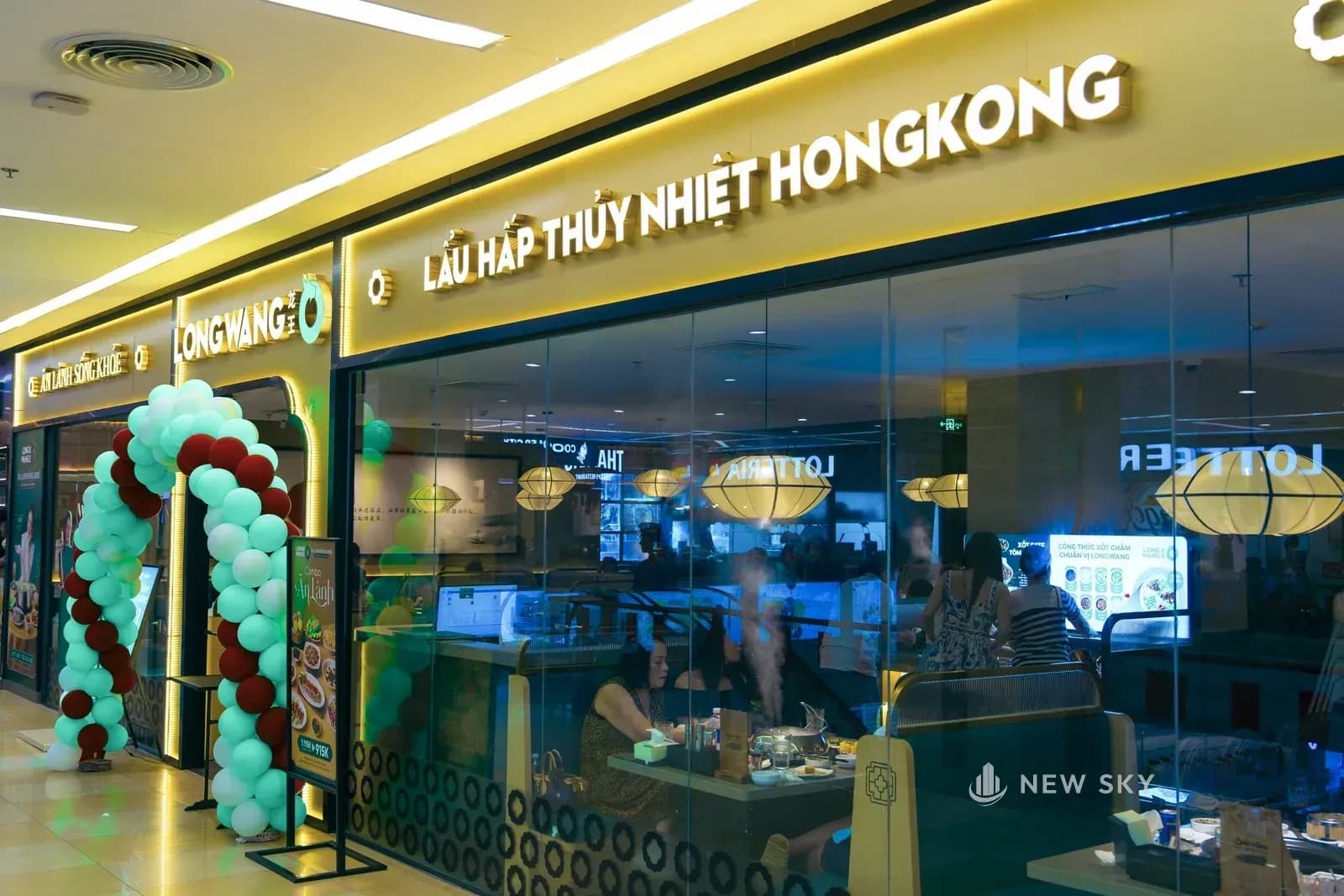 Mặt tiền nhà hàng nhìn từ lối đi trong trung tâm thương mại: dải biển ngang sáng đèn ghi tên thương hiệu và dòng chữ lẩu hấp thủy nhiệt, mặt kính lớn nhìn vào phòng ăn, cạnh cửa dựng một cổng bóng bay.
