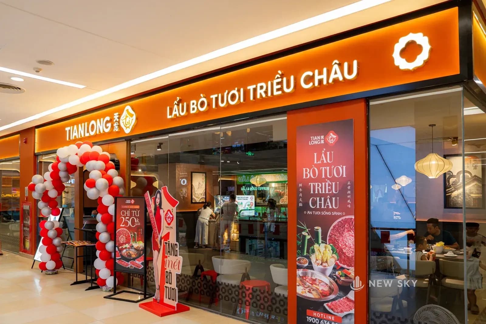 Mặt tiền nhà hàng nhìn từ lối đi trong trung tâm thương mại: dải biển màu cam chạy ngang ghi tên thương hiệu và dòng chữ lẩu bò tươi Triều Châu, dưới là vách kính và một cổng bóng bay dựng cạnh cửa.