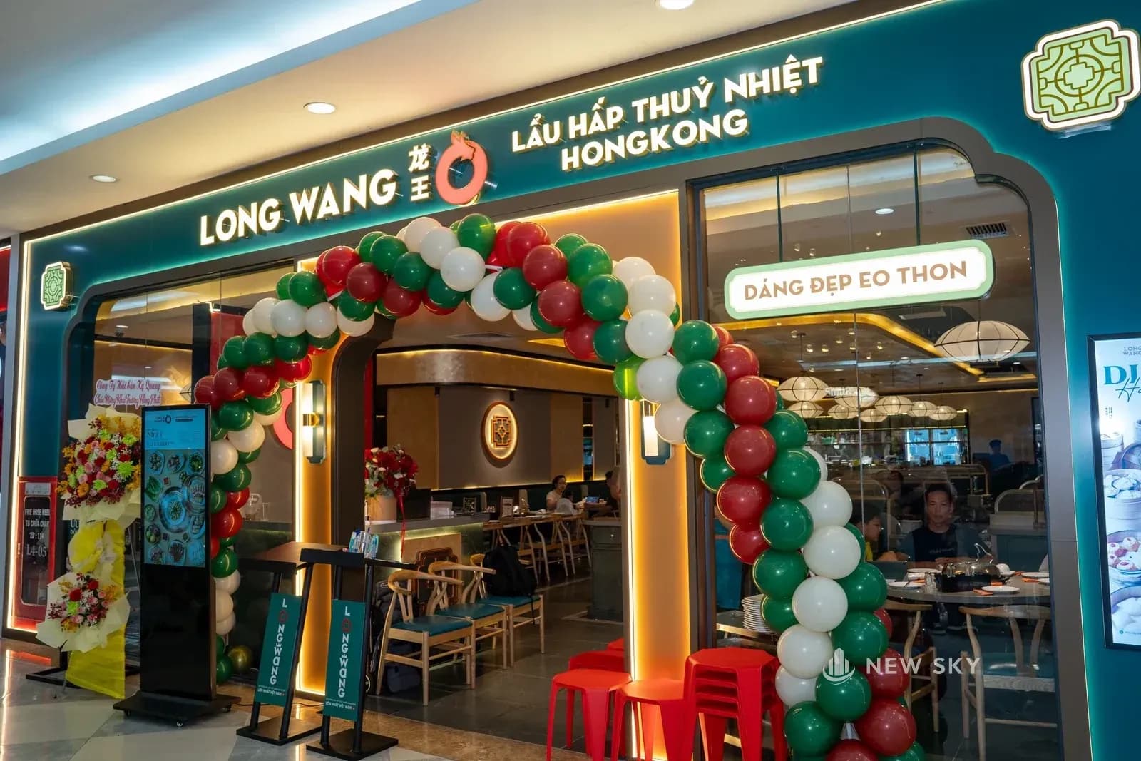 Mặt tiền nhà hàng nhìn từ lối đi trong trung tâm thương mại: cổng vòm với dải biển ngang mang tên thương hiệu và dòng chữ lẩu hấp thủy nhiệt, cạnh cửa dựng một cổng bóng bay.