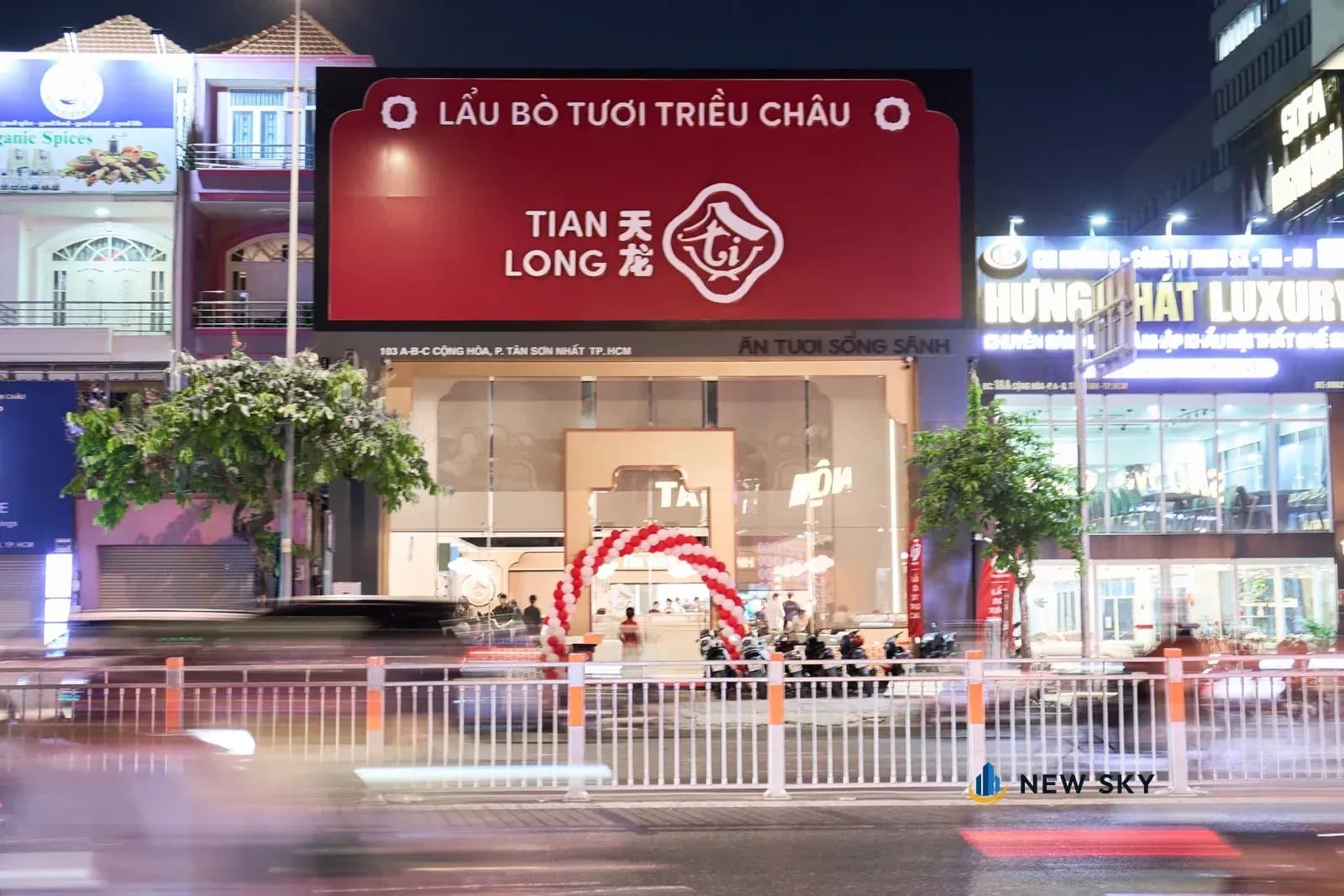 Cùng mặt tiền chụp ban đêm: tấm biển đỏ phát sáng, bên trong nhà đèn vàng hắt ra qua mặt kính, xe chạy qua kéo thành vệt đèn.