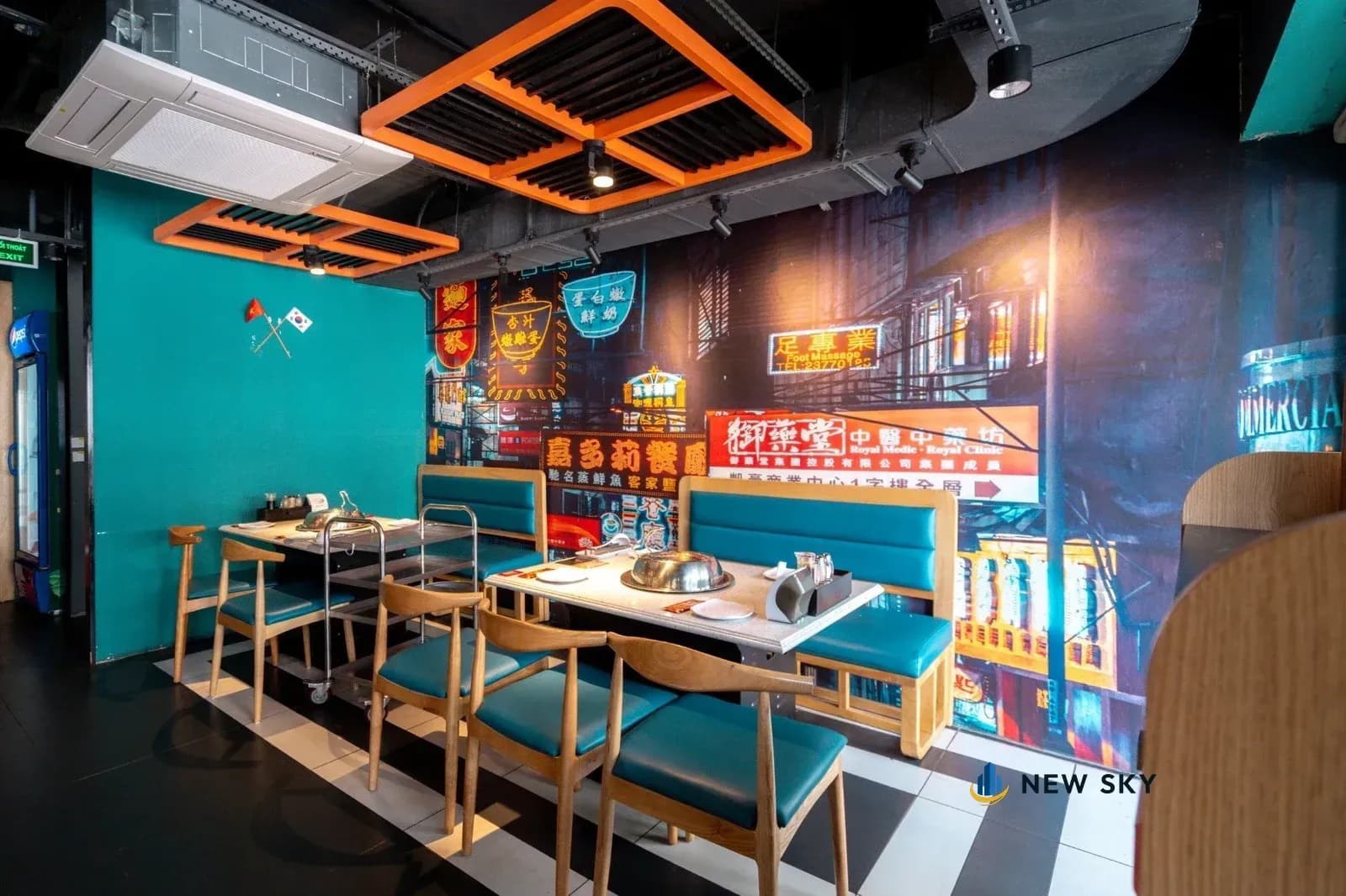 Mảng tường dán ảnh in cỡ lớn chụp một dãy phố về đêm đầy biển hiệu chữ Hoa, kề bên là mảng tường sơn xanh mòng két trơn; bên dưới là khoang ghế băng và bàn có nồi hấp nắp inox.