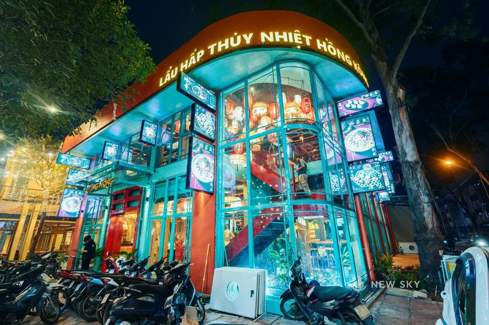 Mặt tiền nhà hàng về đêm: góc nhà bo tròn, hai tầng quây kính khung thép xanh mòng két, phía trên là dải biển cong màu cam đỏ với hàng chữ có đèn; trên các trụ treo những hộp đèn in ảnh món ăn, sau lớp kính góc là cầu thang sắt sơn đỏ.
