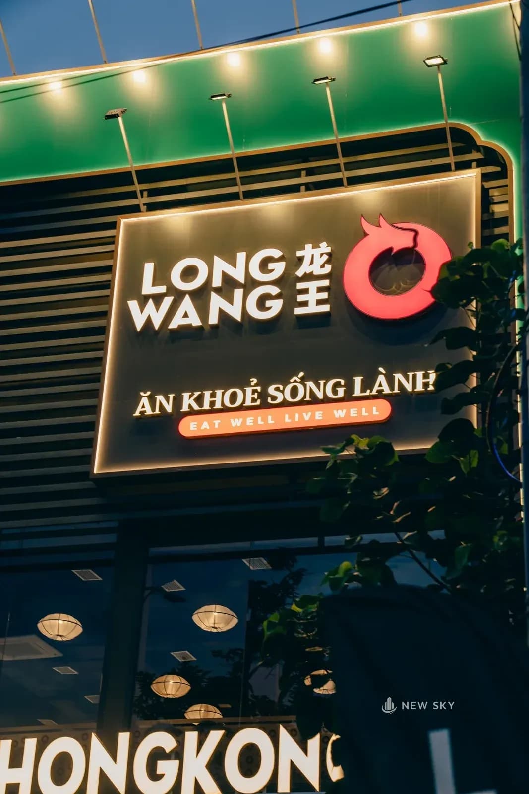 Hộp biển hình chữ nhật viền vàng gắn giữa khối tường xanh ngọc tầng hai, bên trong là tên thương hiệu và một dòng chữ nhỏ; phía dưới thấy một phần hàng chữ nổi màu trắng.