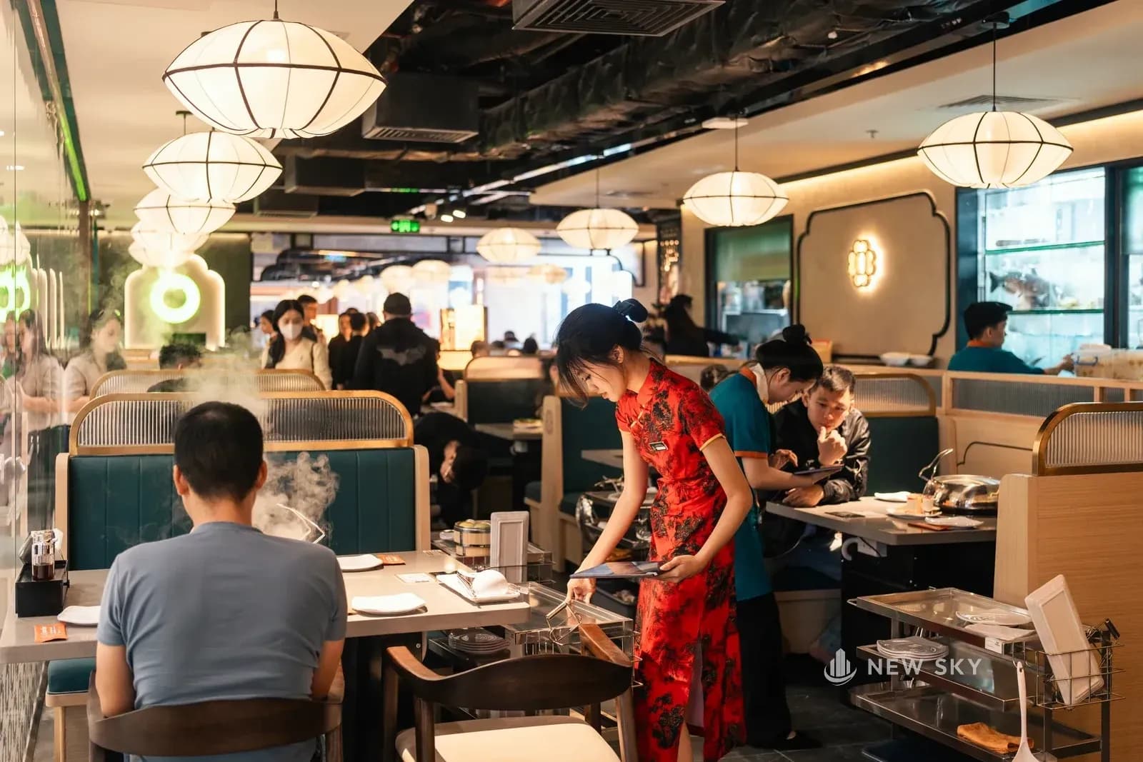 Phòng ăn lúc đông khách: người ngồi trong các khoang ghế băng, nhân viên đứng phục vụ bên bàn, hơi trắng bốc lên từ nồi trên mặt bàn.