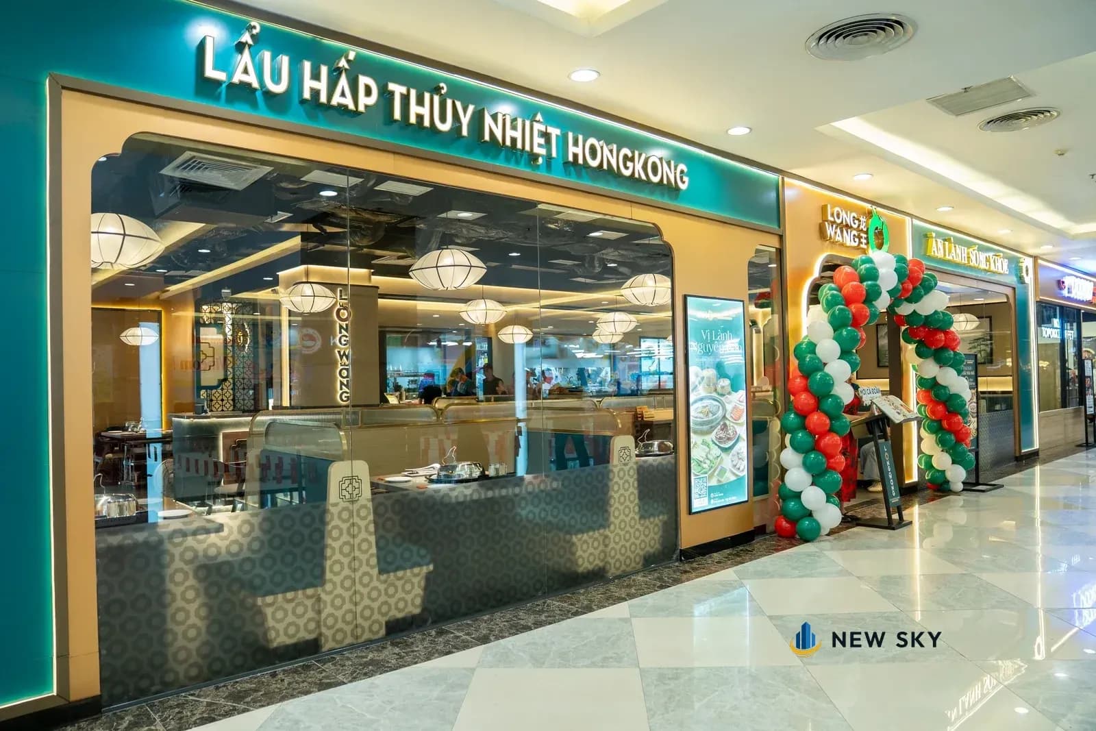 Mặt tiền nhà hàng nhìn từ lối đi trong trung tâm thương mại: dải biển ngang màu xanh ngọc ghi tên món, mặt kính lớn nhìn vào phòng ăn, chân tường ốp gạch hoa văn, cạnh cửa dựng một cổng bóng bay.