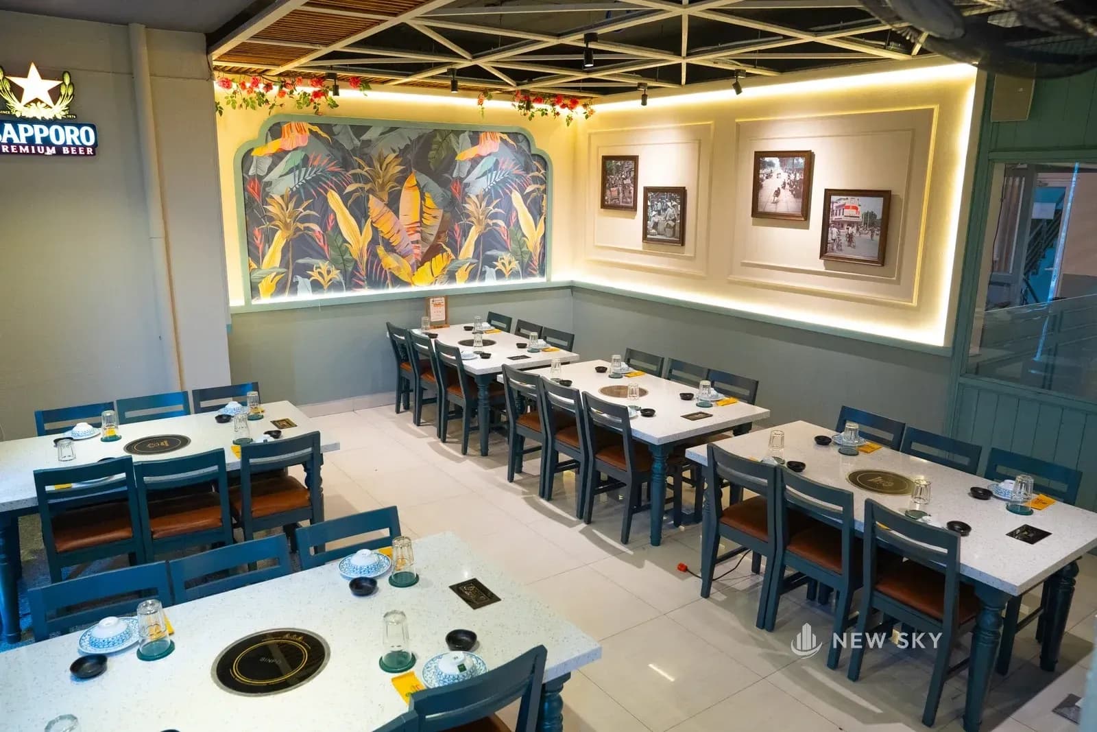 Bức tranh cây lá nhiệt đới khổ lớn trong khung vòm có đèn viền trên mảng tường màu kem, cạnh đó là mấy khung ảnh nhỏ; bên dưới là dãy bàn dài.