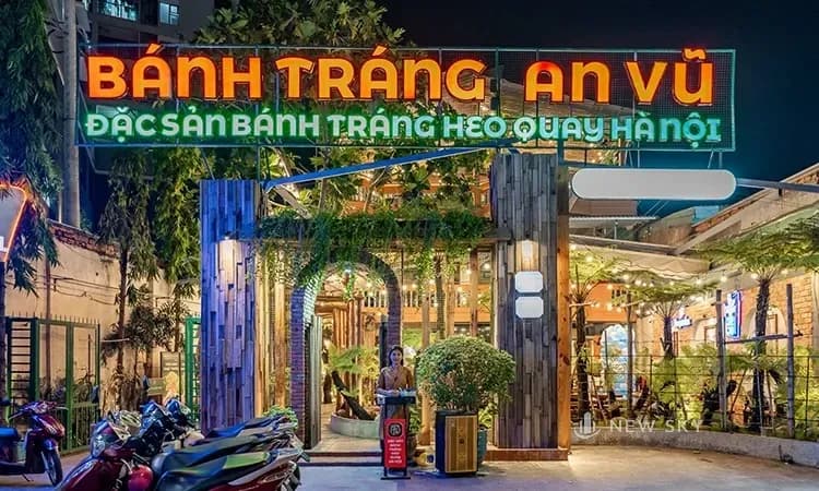 Biển hiệu Bánh Tráng An Vũ chữ nổi phát sáng trên nền xanh, bên dưới là cổng vào ốp gỗ phủ dây leo, chụp ban đêm.