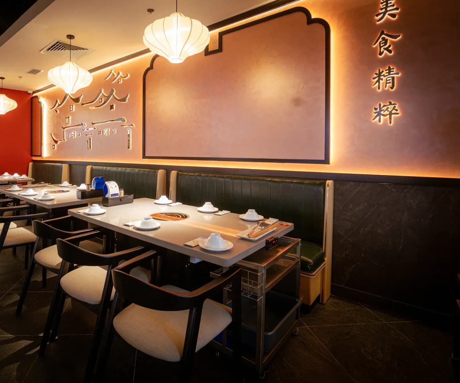 Một góc phòng ăn Tian Long Vincom Royal City: tường sơn hiệu ứng tông cam có khung viền đen uốn hai bậc ở đỉnh, hàng chữ Hán dọc và chữ kim loại hình mái nhà hắt đèn, đèn lồng vải trắng treo trên bàn, ghế băng da xanh và ghế gỗ đen.
