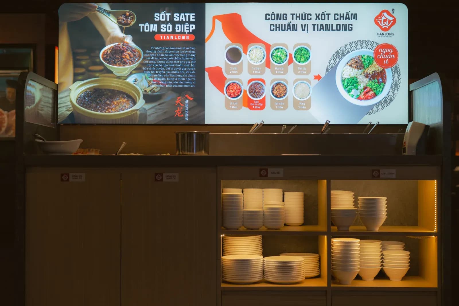 Quầy nước chấm tự chọn ở Tian Long Vincom Plaza Thanh Hoá: bảng đèn hướng dẫn pha xốt phía trên, dãy khay inox đựng xốt trên mặt quầy, tủ dưới chia ngăn xếp sẵn bát và đĩa trắng.