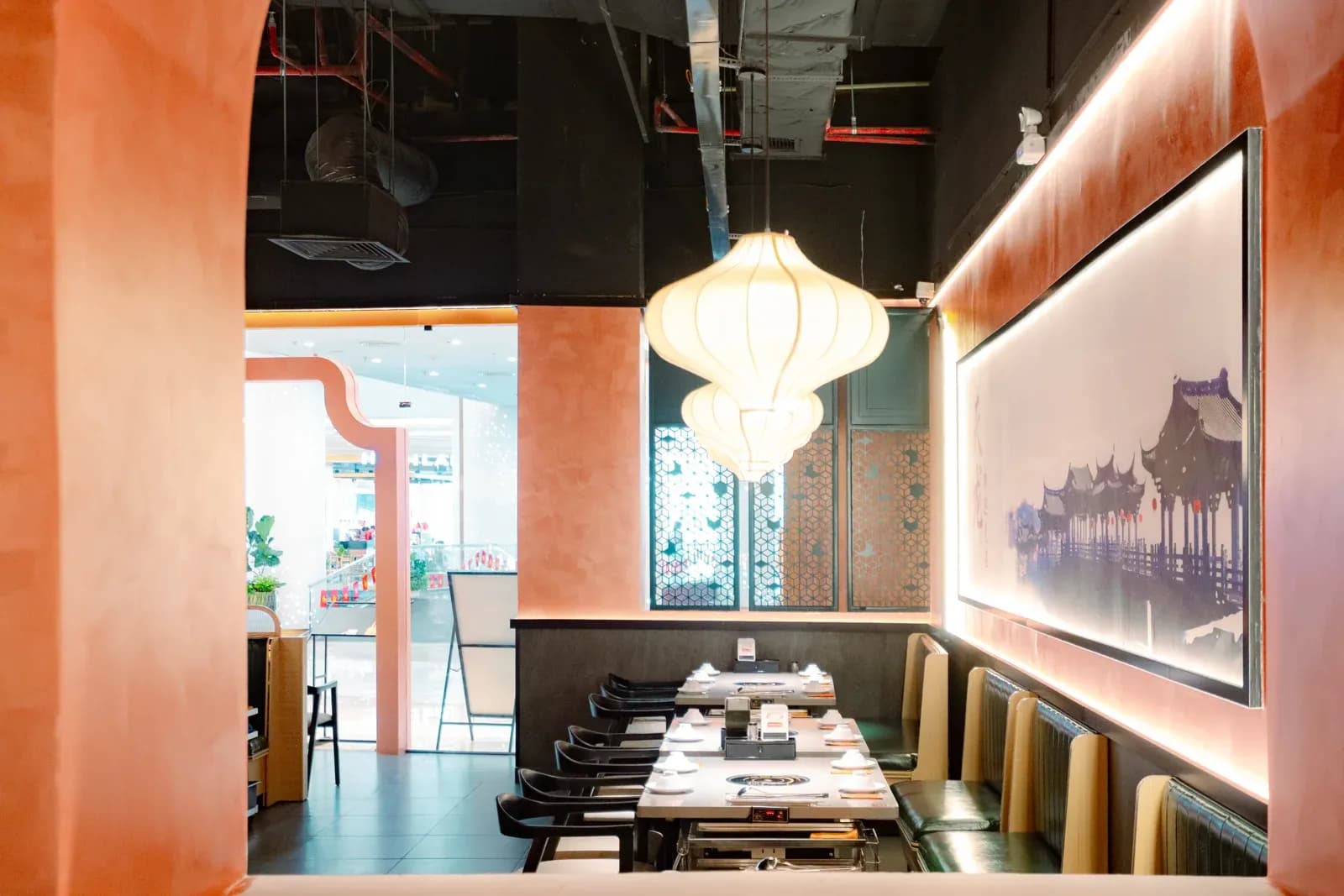 Nhìn từ cổng vòm vào một dãy bàn Tian Long Vincom Mega Mall Smart City: tường sơn cam, mảng tường lớn treo tranh mái đình cổ có đèn hắt, đèn lồng vải treo thấp trên bàn, khoang ghế băng dọc tường.