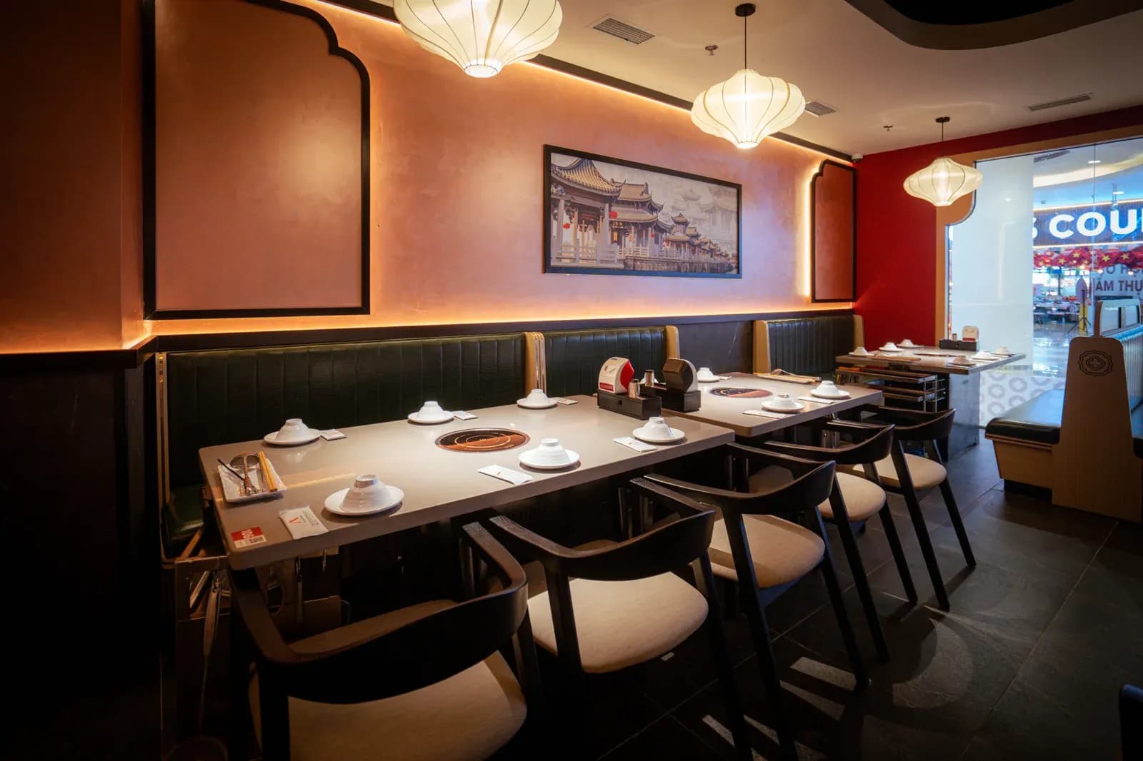 Dãy bàn dài trong Tian Long Vincom Royal City: bàn mặt đá xám có bếp âm, ghế băng da xanh dọc tường, ghế gỗ đen tay cong phía ngoài, tranh phố cổ khung đen trên tường cam, cuối phòng là mặt kính ra hành lang trung tâm thương mại.