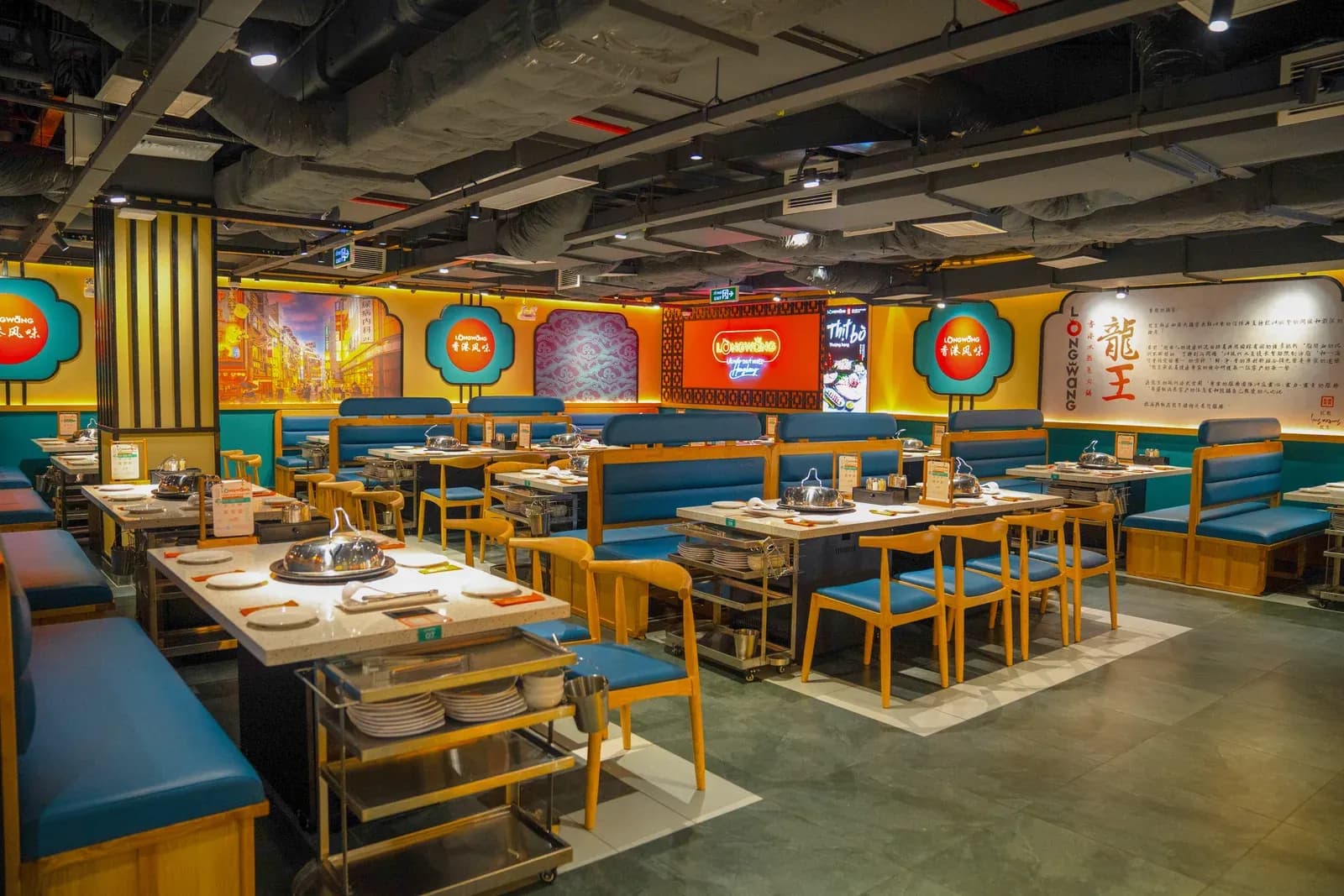 Phòng ăn Long Wang Vincom Center 3/2: tường vàng cam, logo tròn đỏ chữ 香港风味 trong khung hoa viền xanh ngọc, bảng chữ lớn 龍王, biển neon LONGWANG, khoang ghế băng bọc xanh dương, ghế gỗ sáng đệm xanh, bàn mặt đá trắng hạt có nồi hấp, xe đẩy inox cạnh bàn, trần để lộ ống kỹ thuật.