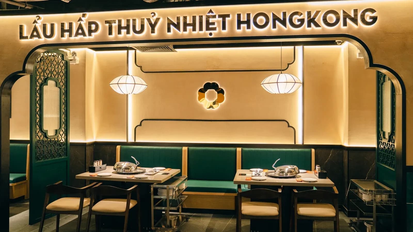 Một khoang ăn Long Wang Vincom 216 Võ Văn Ngân: chữ nổi Lẩu hấp thủy nhiệt Hongkong trên cổng viền đen lượn bậc, tường sơn kem có logo hoa màu đồng hắt sáng, 2 đèn lồng vải trắng, ghế băng xanh lục sẫm, ghế gỗ nâu đệm kem, vách song xanh lục hai bên, xe đẩy inox cạnh bàn.