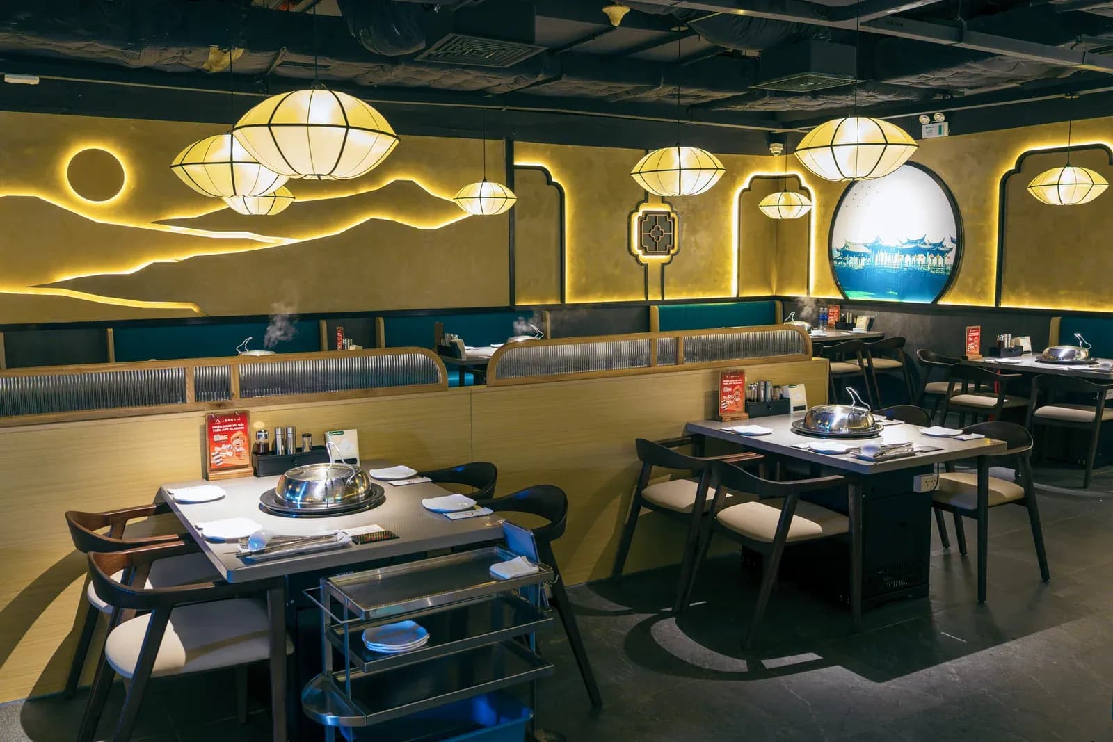 Phòng ăn Long Wang Vincom Plaza Thanh Hoá: tường sơn vàng có đường núi nổi và vầng trăng tròn hắt sáng, tranh tròn khung đen vẽ nhà mái cong bên hồ, đèn lồng vải trắng treo dưới trần đen, dãy ghế băng có vách kính sọc, bàn mặt xám đặt nồi hấp, ghế gỗ nâu đậm đệm kem, xe đẩy inox cạnh bàn.