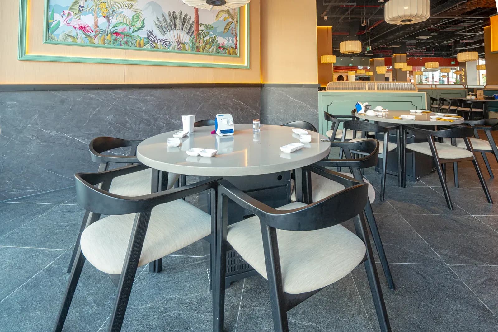 Một góc Bò Tơ Quán Mộc Vincom Mega Mall Royal Island Vũ Yên: bàn tròn mặt đá xám, ghế gỗ sơn đen tay vịn cong đệm vải be, chân tường ốp đá xám vân trắng, tranh nhiệt đới có hồng hạc trong khung xanh, đèn lồng vải trắng treo trần, phía sau là dãy khoang ghế băng.