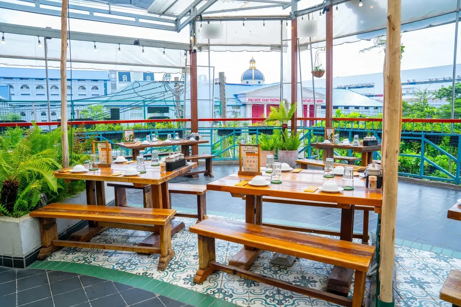 Sân thượng Bò Tơ Quán Mộc 101A-101B Võ Oanh: bàn gỗ dài và ghế băng gỗ mộc kê dưới mái bạt trắng căng trên khung, cột gỗ bóc vỏ dựng đứng, bồn cây dương xỉ, sàn gạch bông, lan can sắt, phía xa là nhà cửa của thành phố.