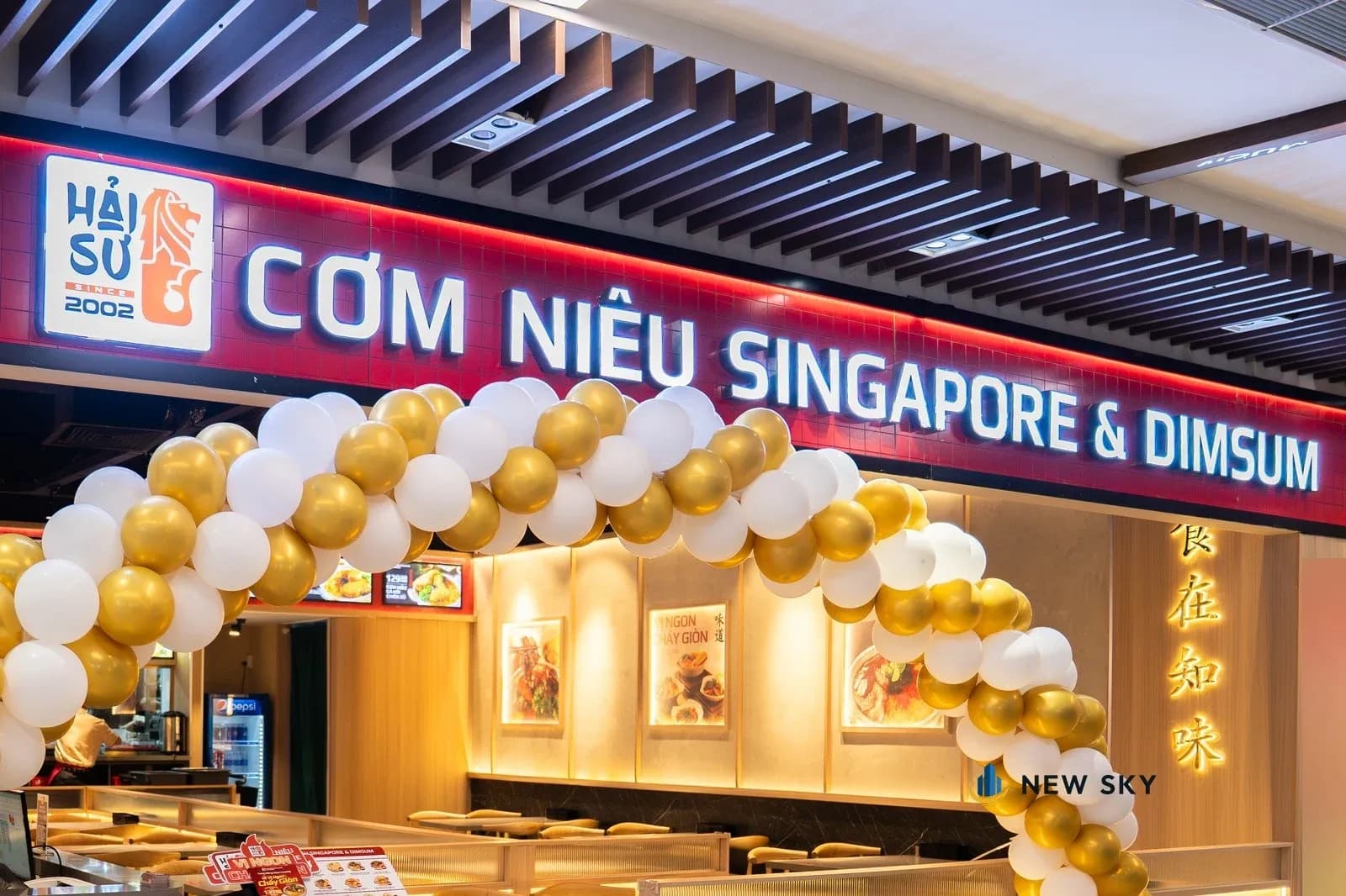 Mặt tiền Cơm Niêu Hải Sư Giga Mall 242 Phạm Văn Đồng: dải biển nền đỏ chạy dài, hộp đèn logo nền trắng hình sư tử biển, chữ nổi hắt sáng; dưới biển là cổng bóng bay vàng trắng, bên trong thấy tường ốp gỗ sáng có tranh ảnh món và chữ Hán hắt sáng.