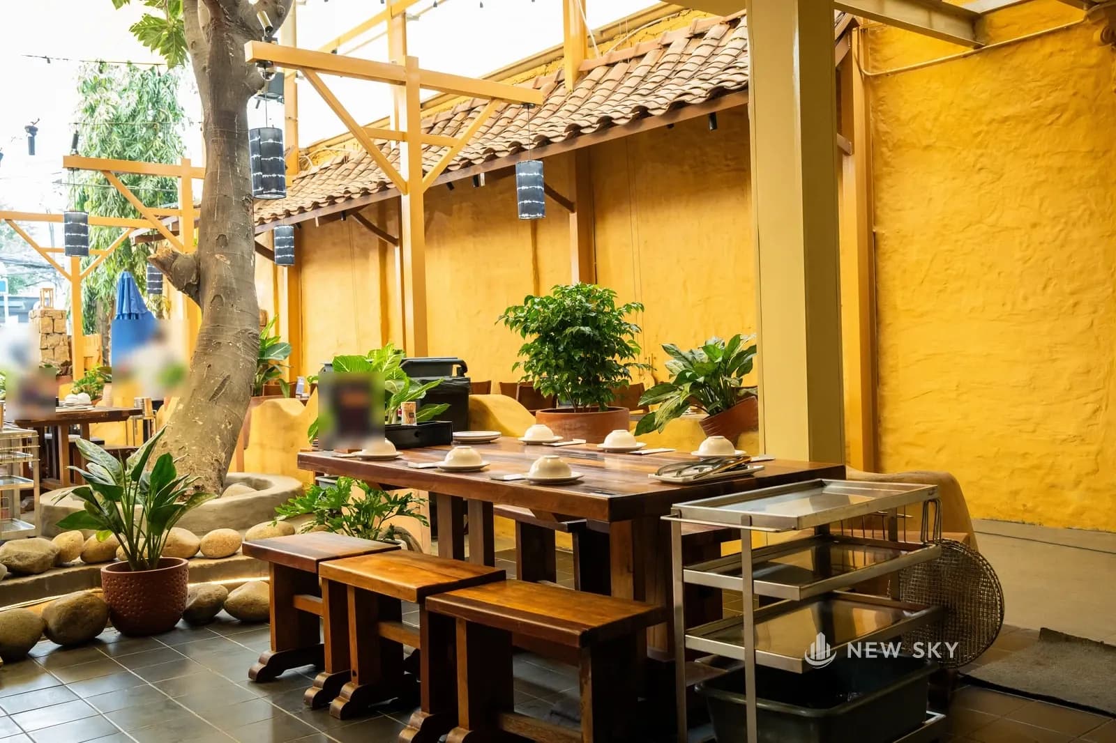 Sân trong Khèn Nướng Sapa số 9 Thoại Ngọc Hầu: thân cây lớn bên trái, dải mái ngói đỏ nâu chạy dọc bức tường trát tay sơn vàng mặt lồi lõm, đèn lồng hình ống màu chàm treo trên khung sơn vàng, một bàn gỗ ghép thanh dày với ghế dài không tựa, xe đẩy inox đứng cạnh bàn, chậu cây và đá cuội ở góc.
