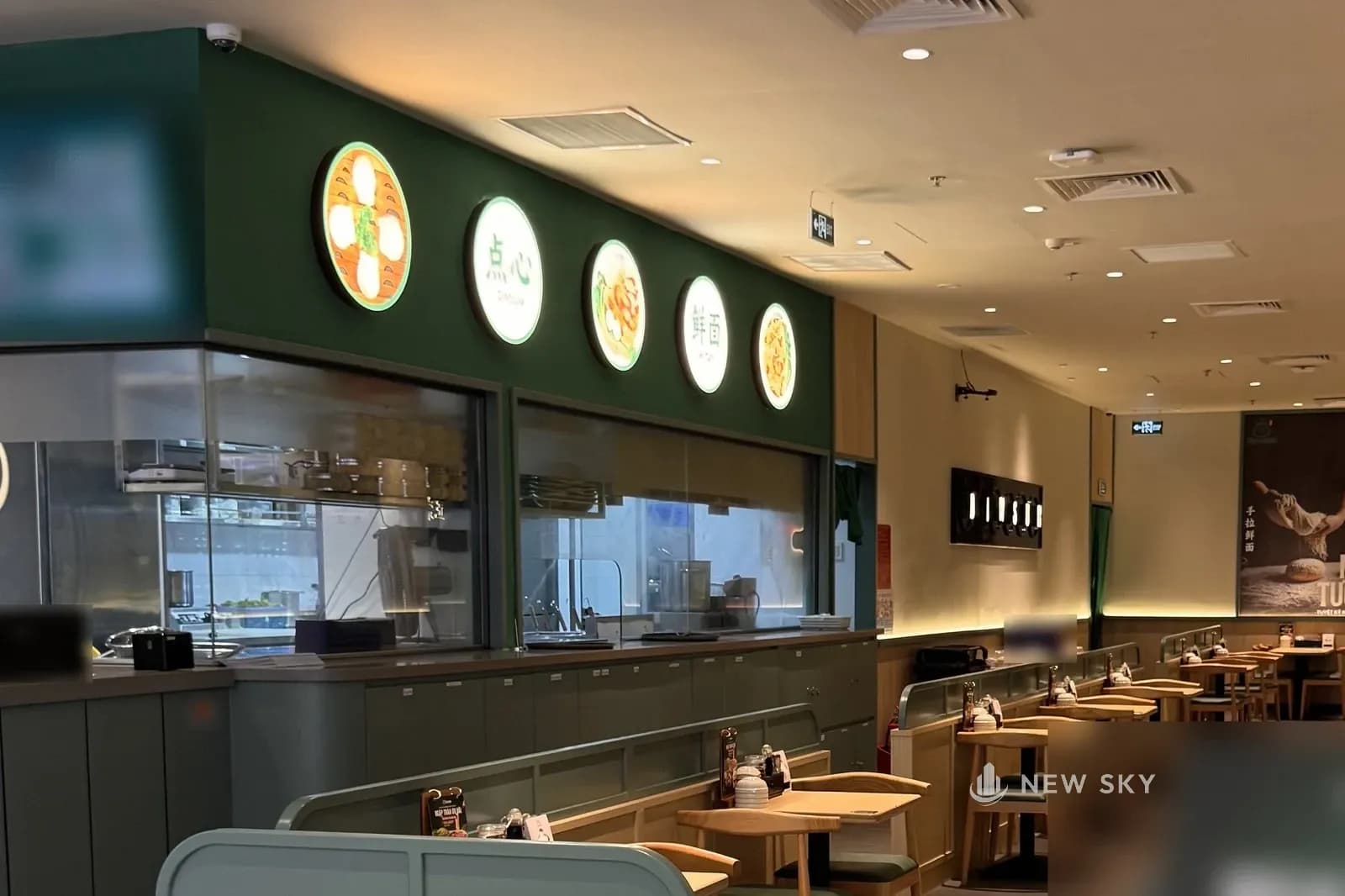 Phòng ăn nhà hàng Cảo Vương tại Vincom Mega Mall Smart City: bên trái là quầy bếp thân xanh lục xám, phía trên là dãy hộp đèn tròn nền kem in hình món xen chữ 点心 và 鲜面, dưới hộp đèn là ô kính nhìn vào bếp; phía trước là bàn gỗ sáng tựa vách thấp bo tròn, cuối phòng là tấm chữ DIMSUM và mép một tranh dọc in món ăn.