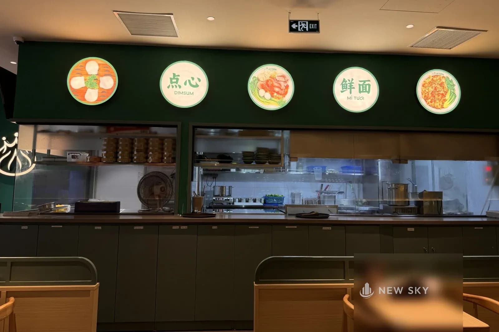 Quầy bếp mở nhà hàng Cảo Vương tại Vincom Mega Mall Smart City: thân quầy xanh lục xám, phía trên là hai ô kính lớn nhìn vào bếp, thấy những chồng xửng tre, giá inox, nồi và bàn inox; trên mảng tường xanh là năm hộp đèn tròn xen hình món với chữ 点心 DIMSUM và 鲜面 MÌ TƯƠI; phía trước là lưng ghế và mặt bàn gỗ sáng.