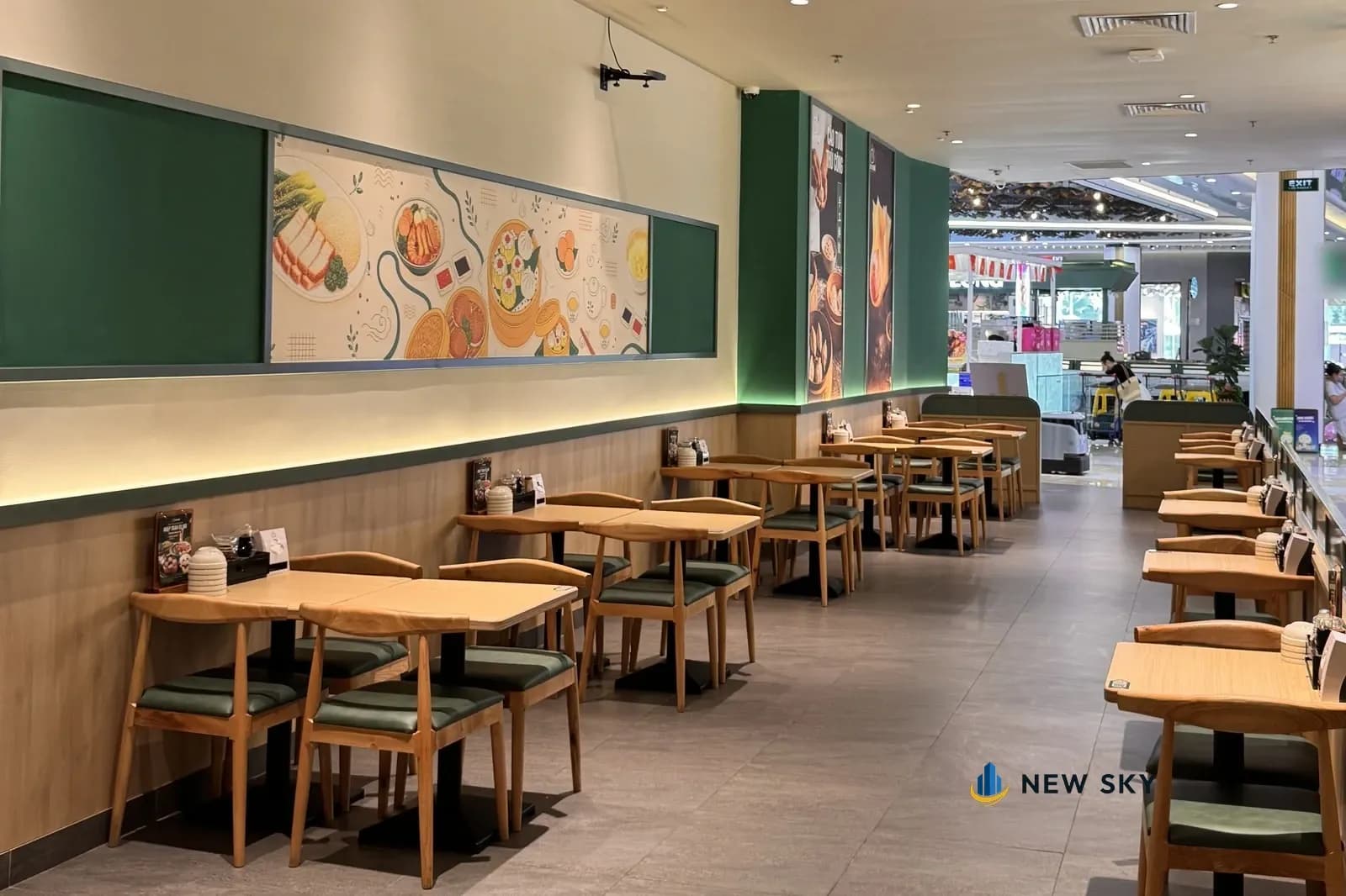 Phòng ăn nhà hàng Cảo Vương tại Vincom Mega Mall Smart City nhìn từ cuối nhà hàng ra lối vào: dãy bàn gỗ sáng ghế đệm xanh sát bức tường có tranh vẽ món trong khung viền xanh và dải đèn hắt chân tường, bên phải là dãy bàn lẻ tựa vách thấp bo tròn, xa hơn là hành lang trung tâm thương mại.