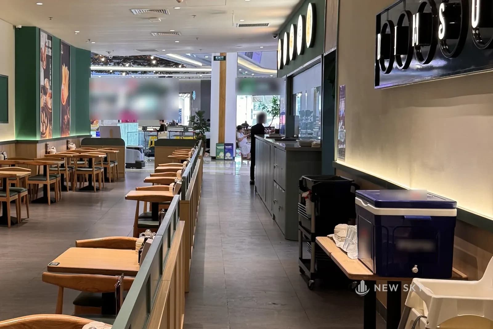 Lối đi dọc quầy trong nhà hàng Cảo Vương tại Vincom Mega Mall Smart City: bên trái là các bàn gỗ sáng tựa vách thấp bo tròn, phía trên quầy bếp là dãy hộp đèn tròn, bên phải là tấm nền đen gắn chữ DIMSUM mỗi chữ trong một vòng tròn sáng, cuối lối là hành lang trung tâm thương mại.