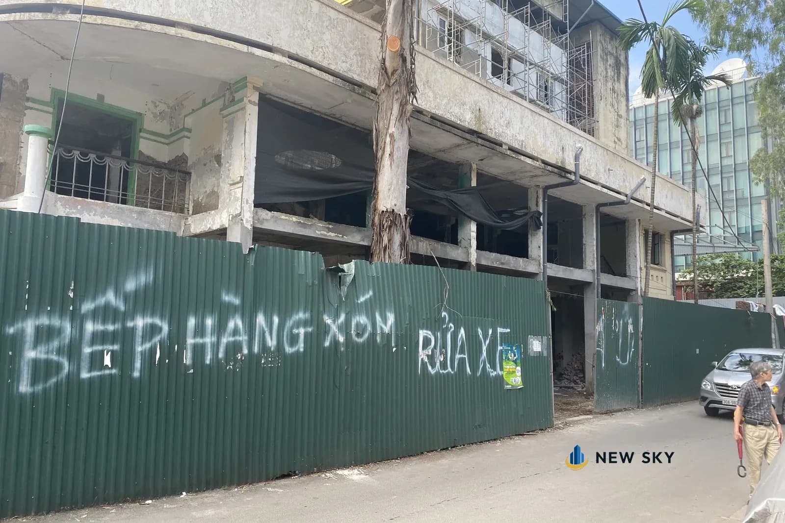 Khối nhà cũ ở 84 Ngọc Khánh lúc khảo sát mặt bằng: góc nhà bo cong, tường loang lổ, các tầng trống trơn, giàn giáo ở tầng trên; bên dưới quây hàng rào tôn xanh có chữ xịt sơn "BẾP HÀNG XÓM", "RỬA XE"; ngoài đường có một người đi bộ và một xe ô tô đỗ.