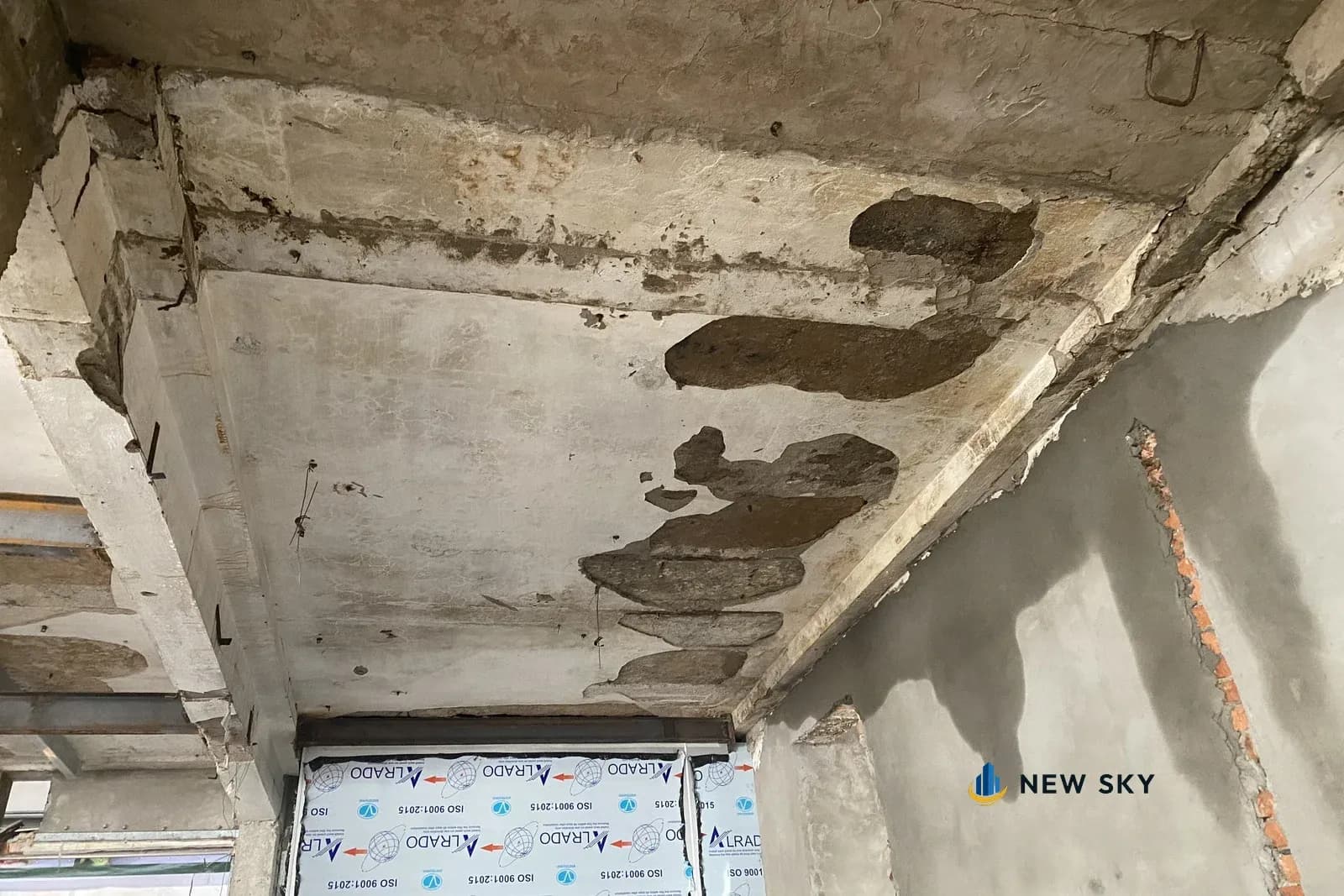 Trần bê tông của nhà cũ bong từng mảng vữa, lộ lớp bên trong; mép dưới là một dầm thép chữ I; bên dưới là mấy tấm khung cửa nhôm còn bọc nilon.