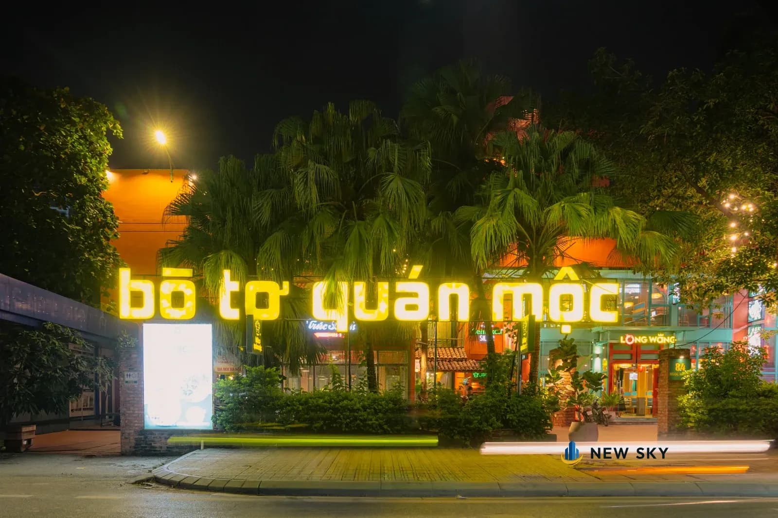Mặt phố 84 Ngọc Khánh về đêm: dãy chữ lớn tên Bò Tơ Quán Mộc phát sáng vàng đặt trên dải cây, phía sau là hàng cọ và khối nhà sơn cam; bên phải là cổng Long Wang có biển tên đèn, cột cổng mang số 84.
