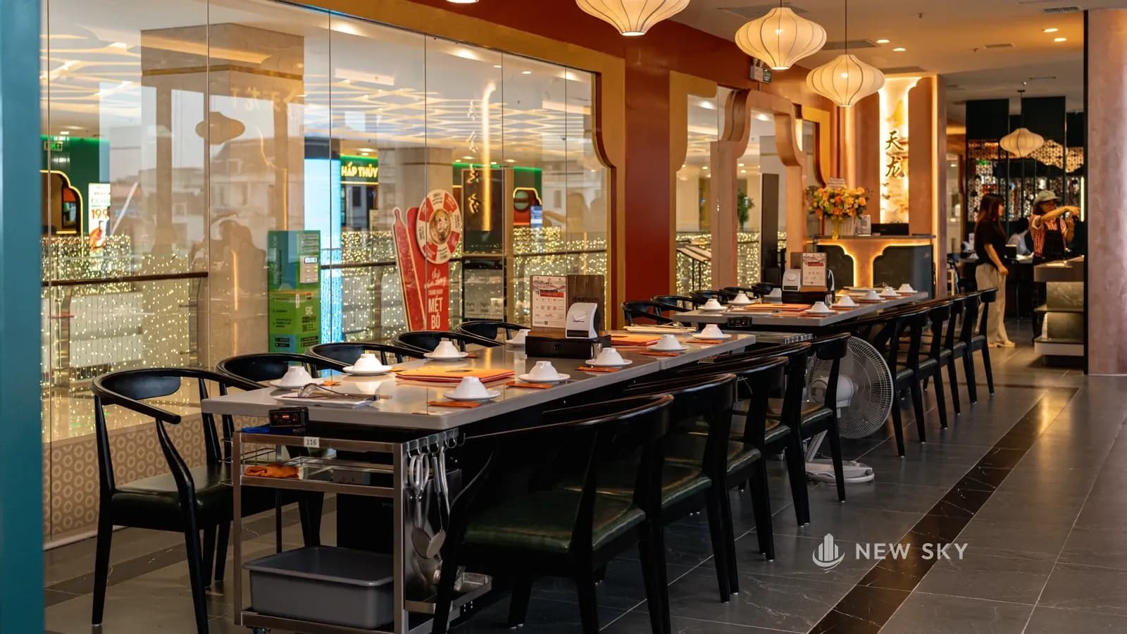 Dãy bàn lẩu ghép dài kê dọc vách kính lớn phía hành lang tầng 3, ghế đen tựa cong hai bên, đèn lồng vải trắng thả phía trên; cuối dãy là một quầy có chữ 天龙 và lẵng hoa.