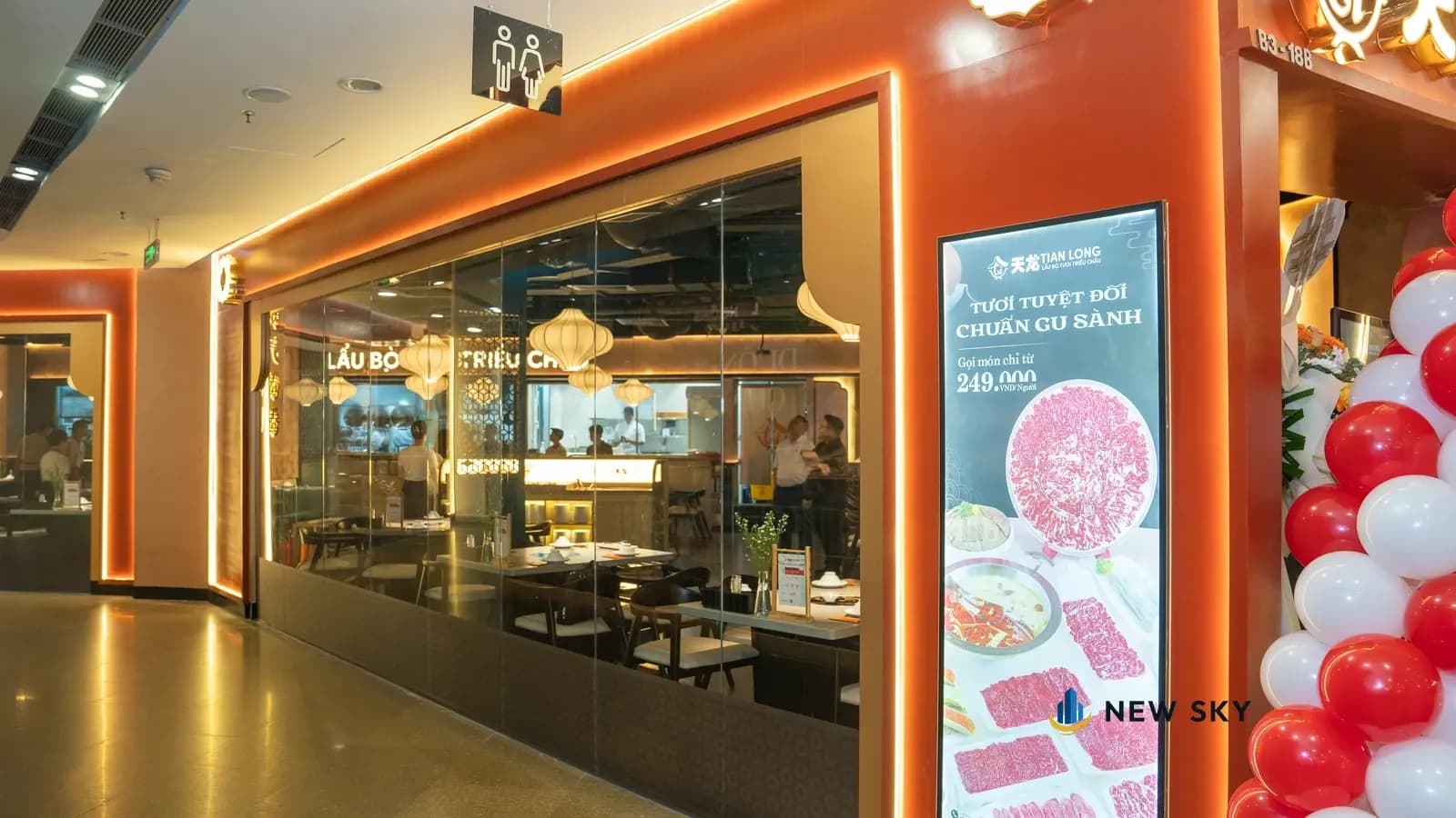 Mặt tiền Tian Long nhìn dọc hành lang tầng hầm B3: khối ốp đỏ cam viền đèn, vách kính dài, màn hình quảng cáo lẩu bò đứng cạnh cửa; qua kính thấy đèn lồng trắng và dải bếp phía trong.