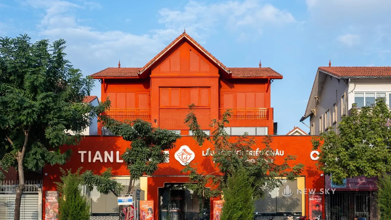 Mặt tiền Tian Long Vũ Trọng Khánh ban ngày: căn nhà mái ngói đỏ, tường sơn đỏ cam, ban công ở tầng trên; phía trước là khối 1 tầng mang dải chữ TIANLONG, logo và "LẨU BÒ TƯƠI TRIỀU CHÂU"; hàng cây vỉa hè che một đoạn chữ; căn bên phải cùng kiểu mái, tường trắng.