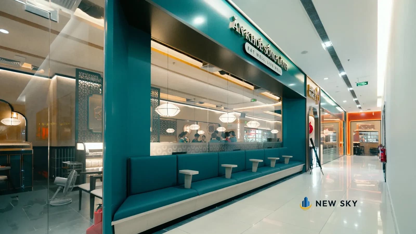 Hốc lùi vào trên mặt tiền Long Wang Vincom 191 Bà Triệu: dãy ghế băng xanh với các mặt bàn nhỏ quay ra hành lang; dải biển xanh đậm chạy ngang bên trên, ghi "ĂN KHOẺ SỐNG LÀNH" và "EAT WELL, LIVE WELL", phía sau là vách kính.