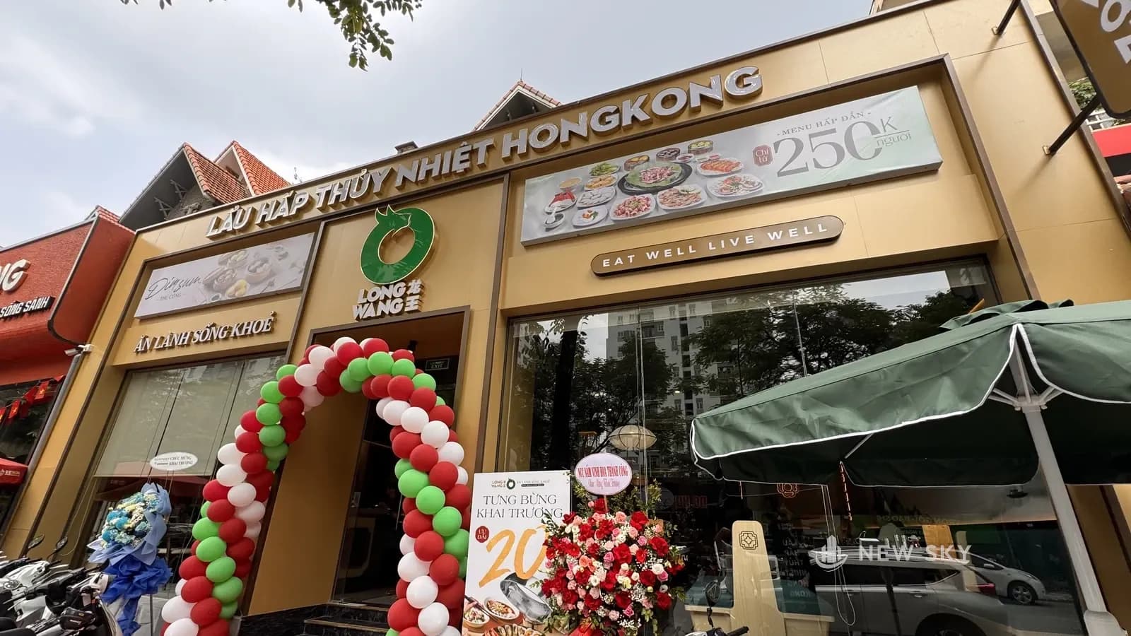 Mặt tiền Long Wang Linh Đàm ngày khai trương, nhìn chéo từ vỉa hè: mặt ốp màu vàng cát, chữ nổi "LẨU HẤP THỦY NHIỆT HONGKONG" chạy ngang mép trên, logo tròn xanh lá trên cửa, ô kính lớn ở tầng trệt, cổng bóng bay đỏ xanh trắng và ô dù xanh trước cửa, mái ngói đỏ của căn biệt thự phía sau.