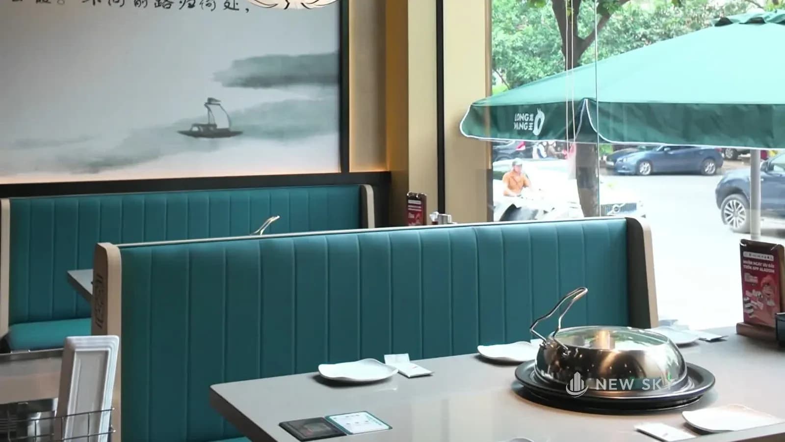 Khoang ghế băng bọc màu xanh ngọc kê sát vách kính mặt tiền, bàn mặt đá xám có nồi hấp inox ở giữa; ngoài kính là ô dù xanh và đường phố.