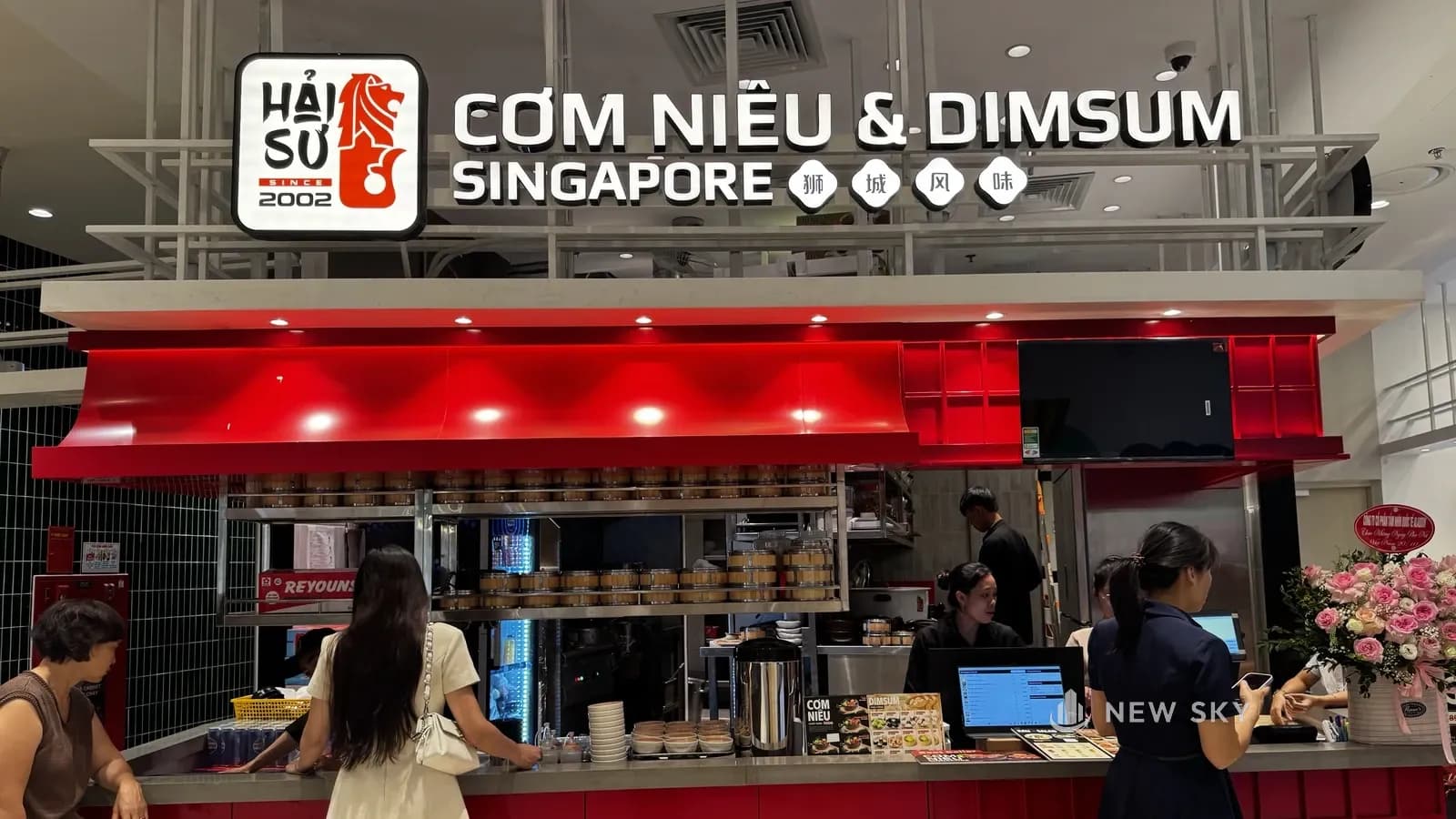 Mặt trước quầy Cơm Niêu Hải Sư nhìn thẳng: hộp đèn logo sư tử biển và chữ nổi "CƠM NIÊU & DIMSUM SINGAPORE" trên khung thép trắng, mái hắt đỏ uốn cong, kệ inox xếp xửng hấp tre, thân quầy đỏ, khách đứng gọi món.
