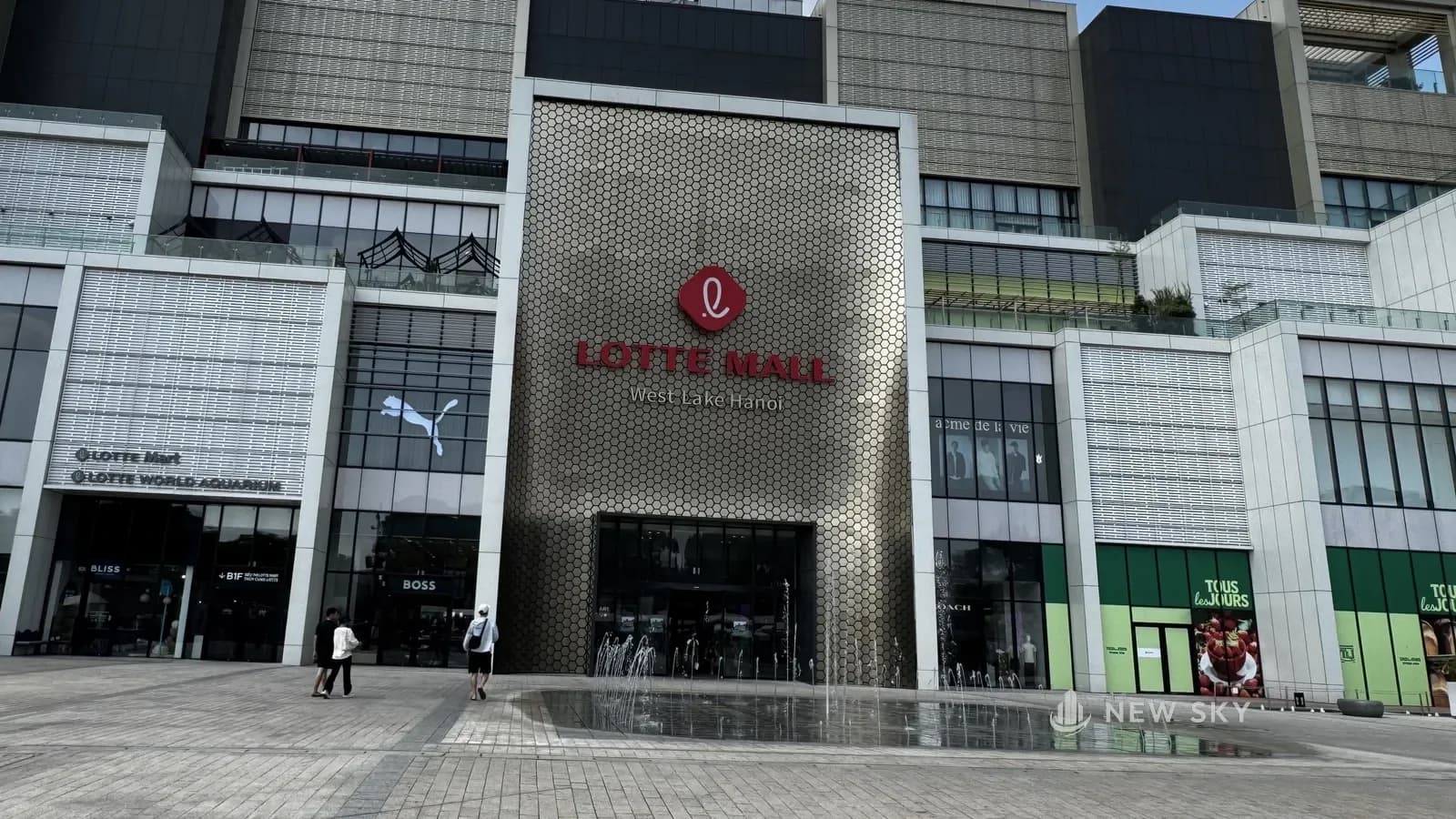 Mặt tiền trung tâm thương mại Lotte Mall West Lake Hanoi: mảng ốp lục giác ánh vàng mang logo và chữ "LOTTE MALL", sân lát đá phía trước có đài phun nước.