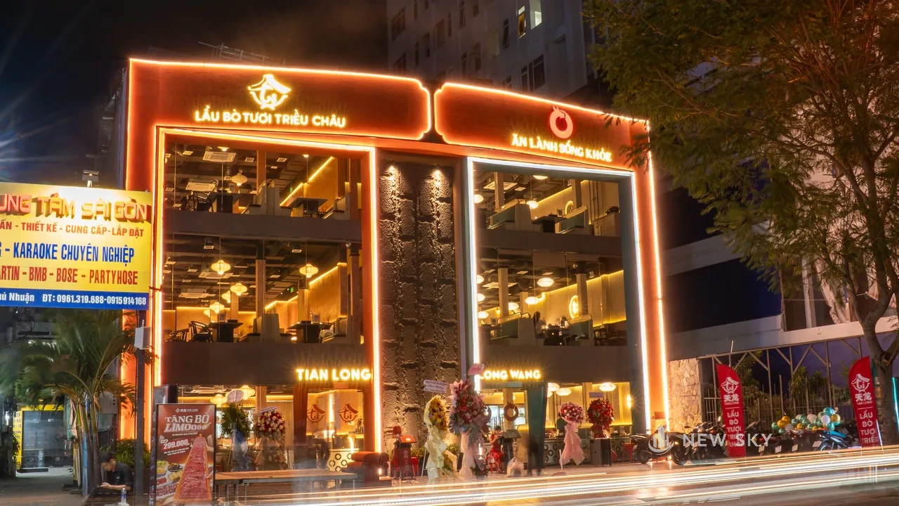 Hai mặt tiền nhà hàng liền nhau buổi tối, viền đèn chạy quanh khung, chữ nổi tên nhà hàng và hộp đèn khẩu hiệu cùng phát sáng, hai cờ dọc dựng bên phải.
