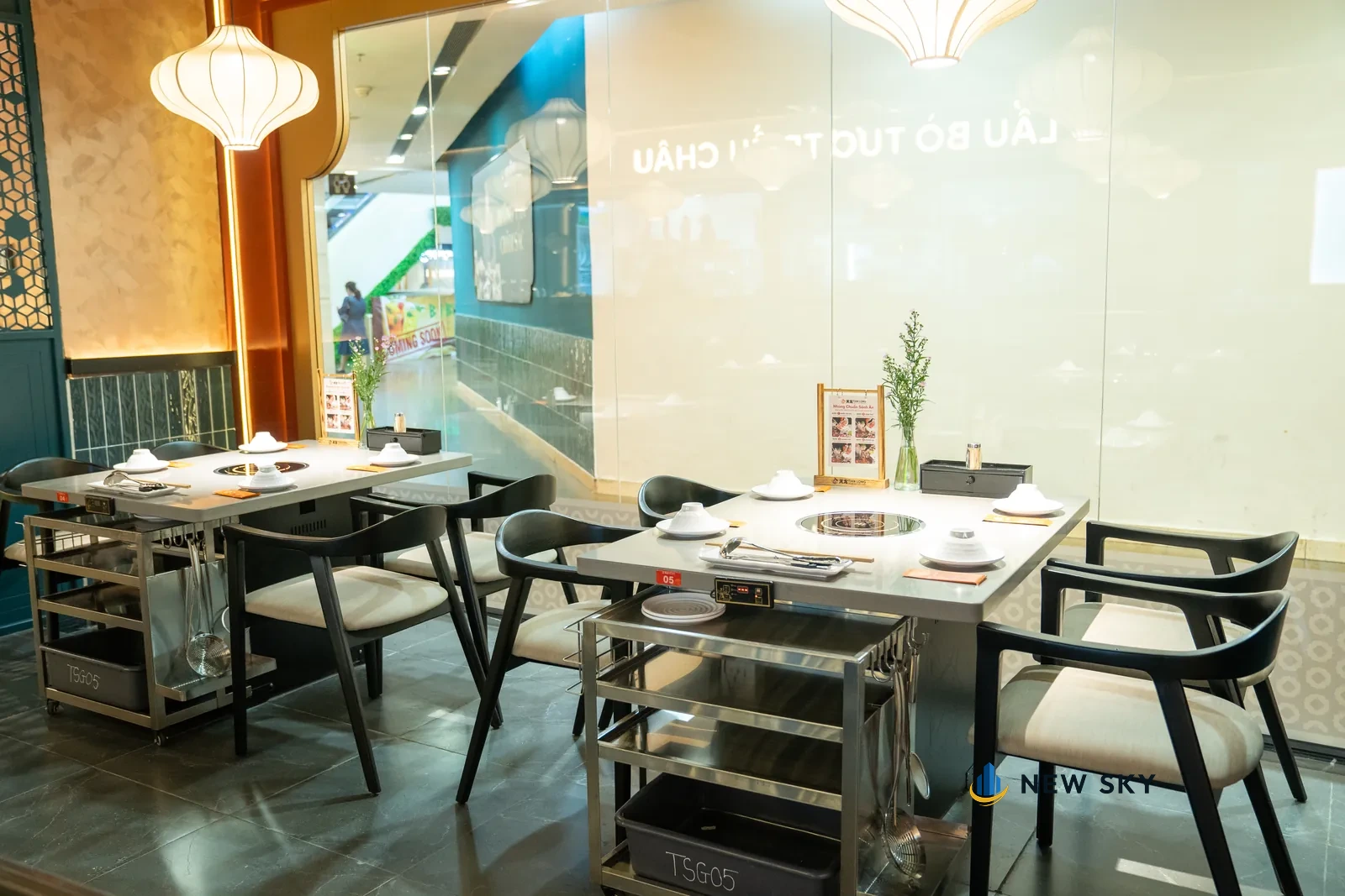 Hai bàn lẩu đặt dọc vách kính, mỗi bàn một bếp âm bàn tròn, xe đẩy inox nhiều tầng đặt cạnh từng bàn, đèn lồng vải trắng treo phía trên.
